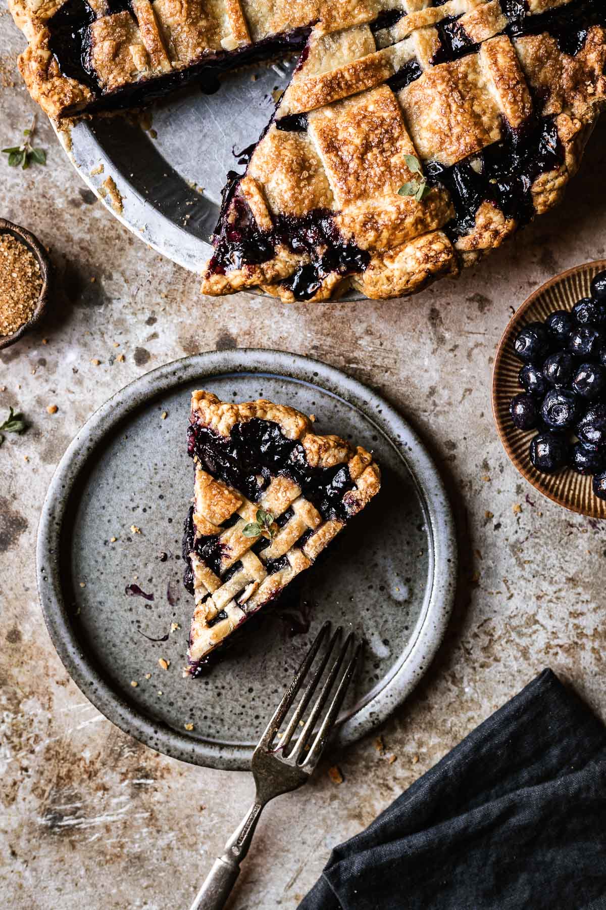 A slice of lattice topped blueberry pie on a grey ceramic plate. The rest of the pie peeks into view at top.