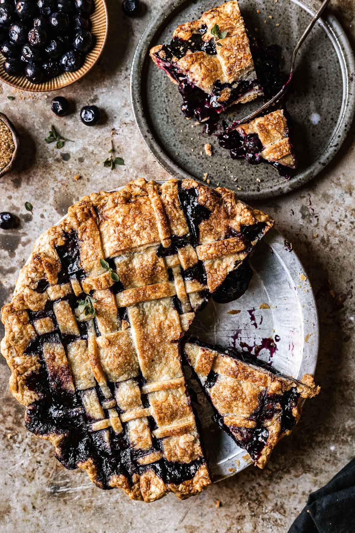 A baked lattice topped blueberry pie with several slices removed from the pan. A single slice sits on a speckled grey plate near the pie pan.