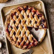 A golden brown lattice topped baked cherry rhubarb pie on a parchment lined baking sheet. A slice has been removed from the pie.