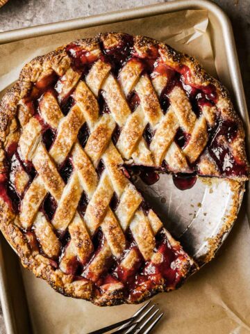 A golden brown lattice topped baked cherry rhubarb pie on a parchment lined baking sheet. A slice has been removed from the pie.