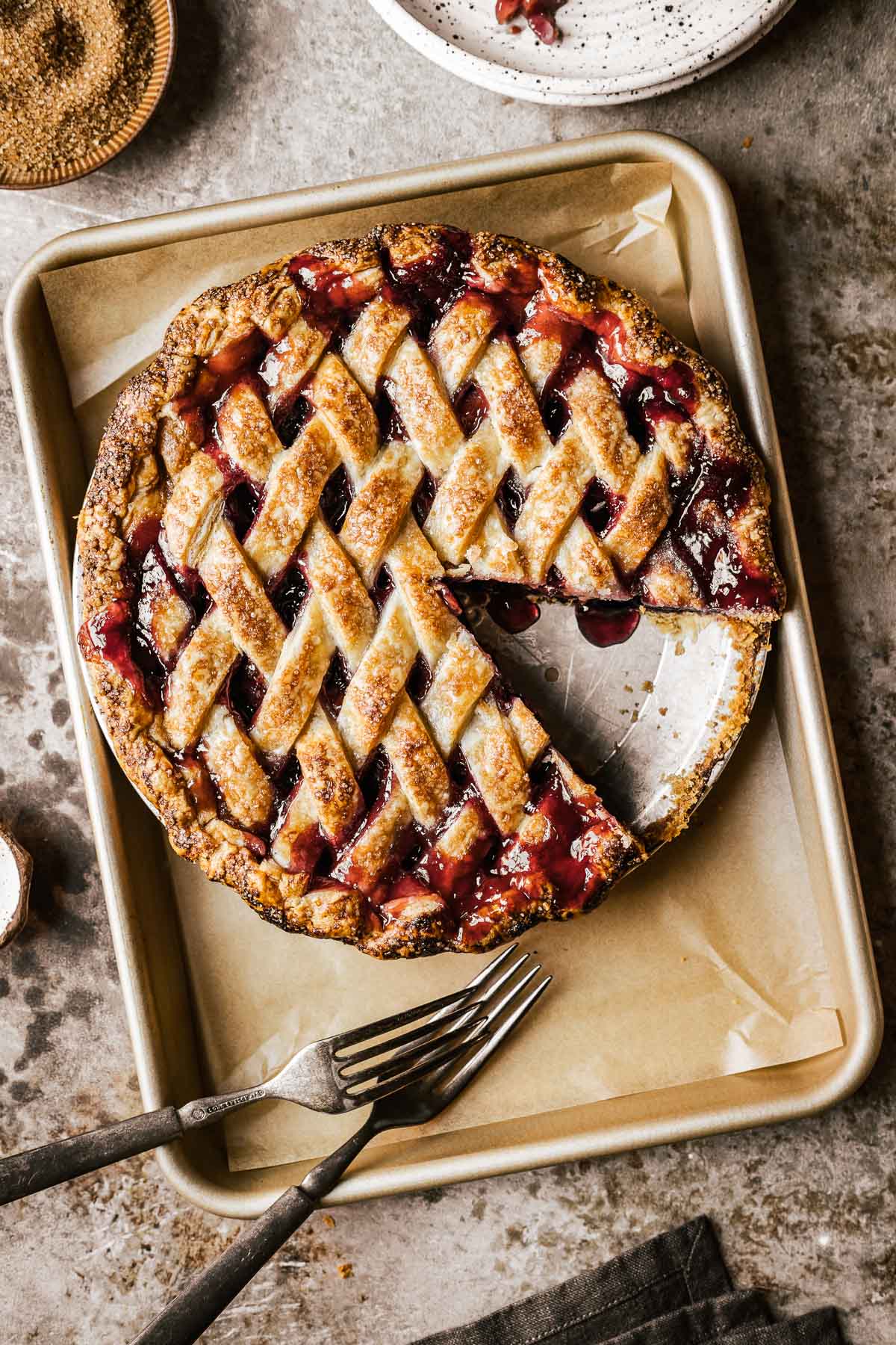 A golden brown lattice topped baked cherry rhubarb pie on a parchment lined baking sheet. A slice has been removed from the pie.