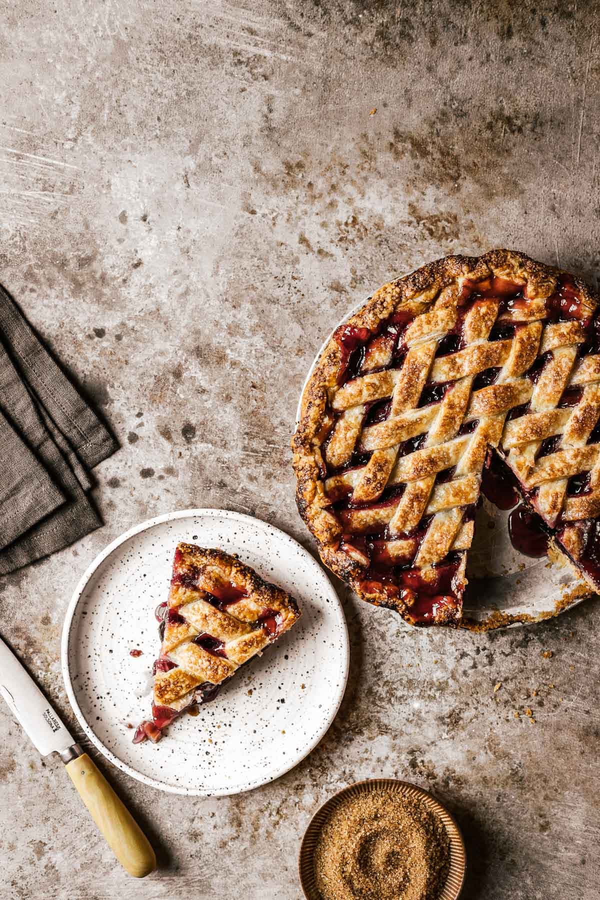 A slice of lattice topped pie on a white ceramic plate with the rest of the pie nearby. A brown napkin, knife and bowl of sugar rest nearby.