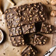 A single layer square chocolate cake on brown parchment paper with nuts sprinkled on top of whipped chocolate ganache frosting.