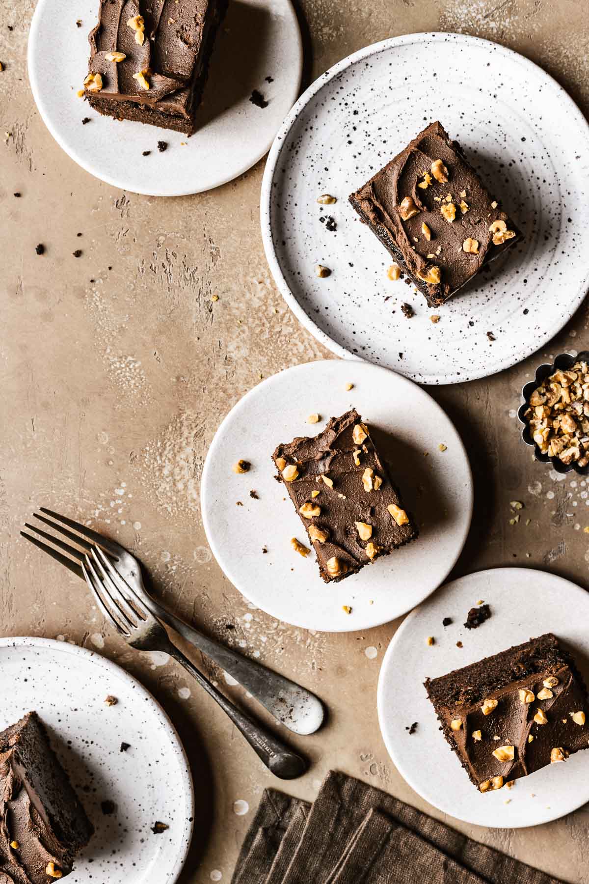 Five slices of chocolate walnut cake on ceramic plates with forks and a brown napkin nearby.
