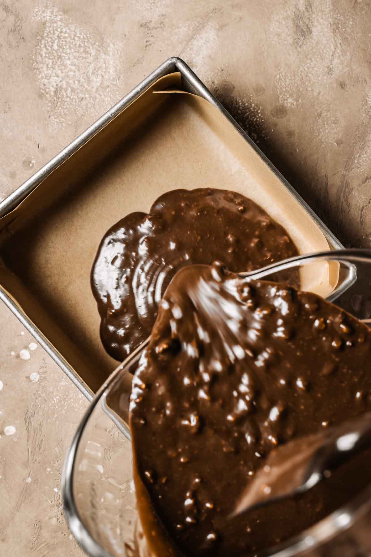 Chocolate walnut cake batter being poured from a glass mixing bowl into a parchment lined square pan.