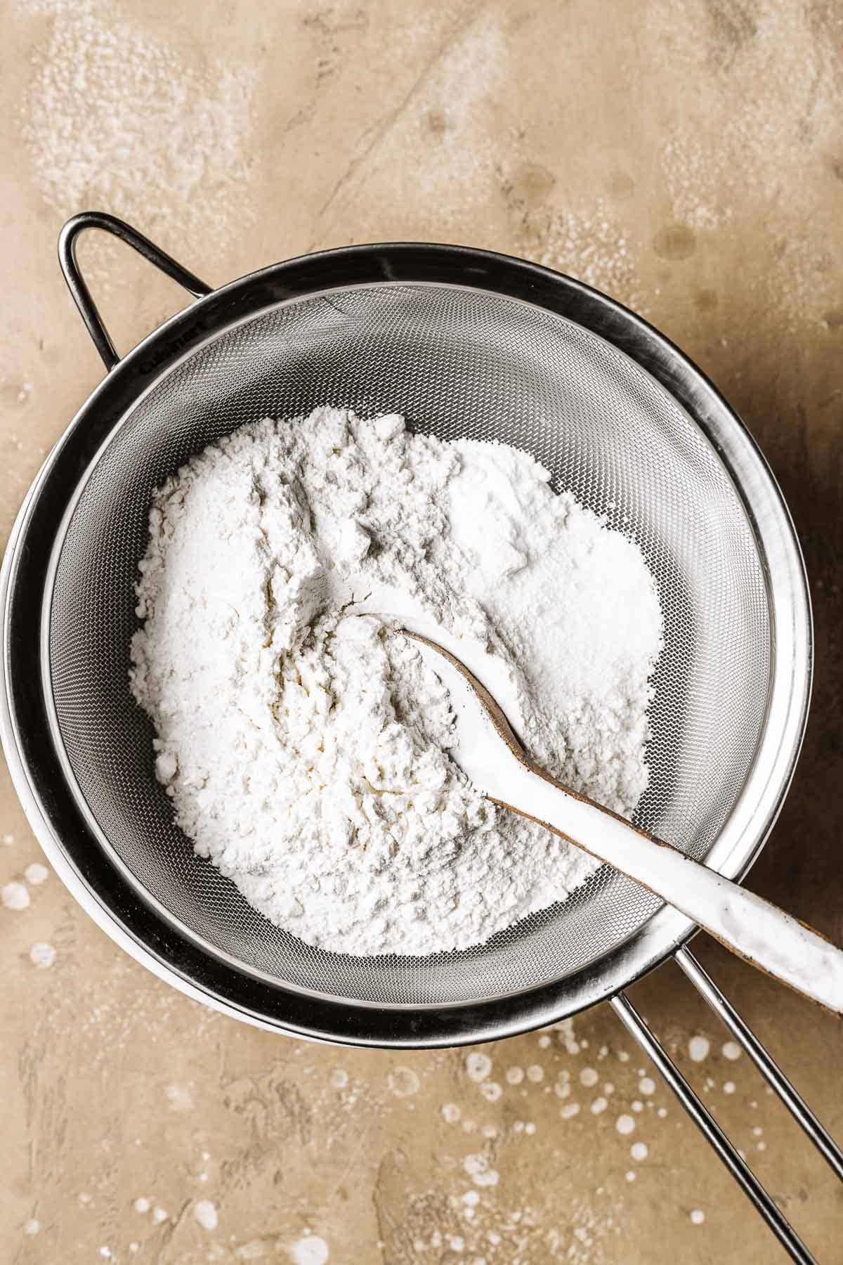 A metal sieve filled with flour, baking soda and salt over a white ceramic bowl. A ceramic spoon rests in the flour mixture.
