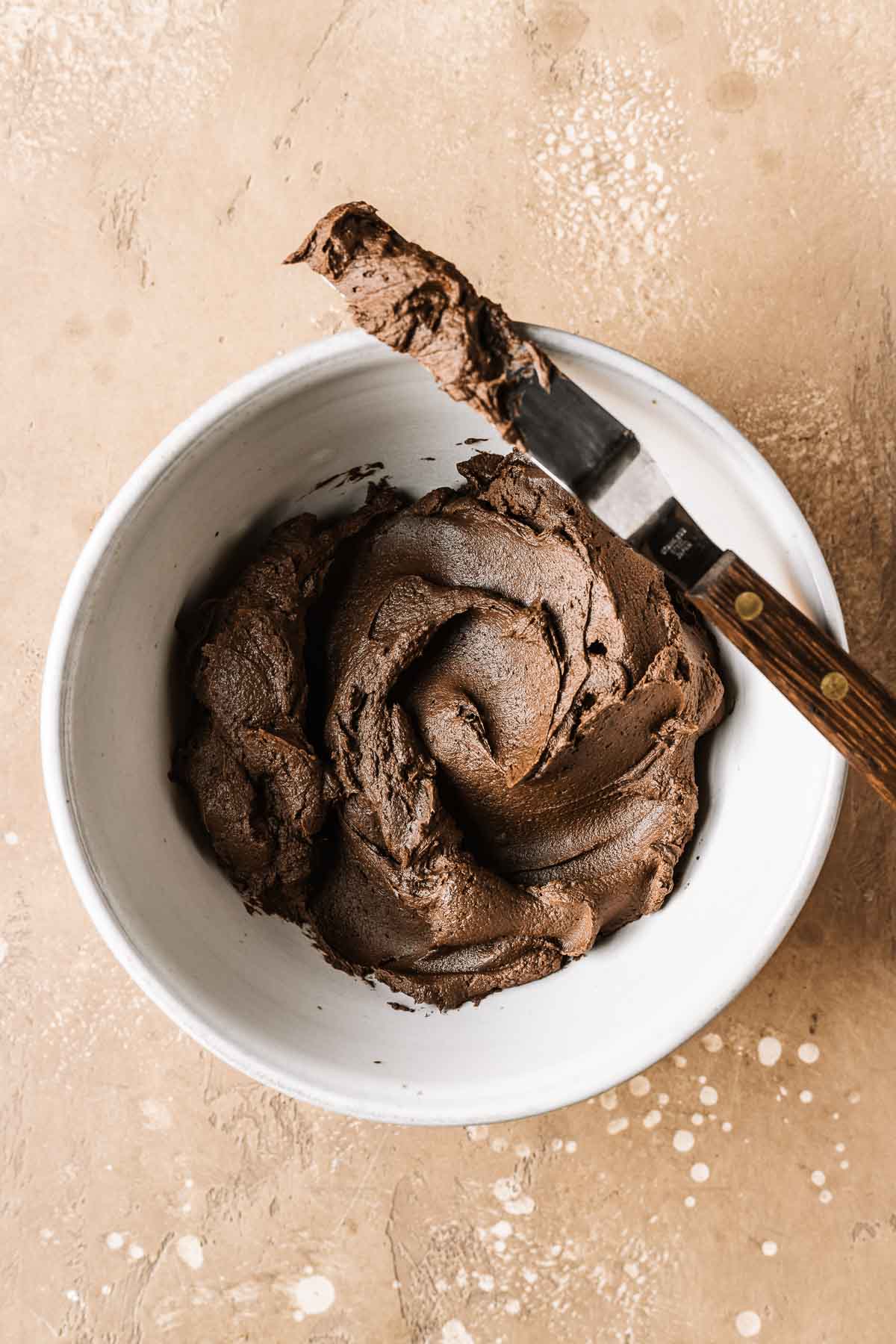 A white ceramic bowl of whipped chocolate ganache with a small metal spatula resting on the edge of the bowl.