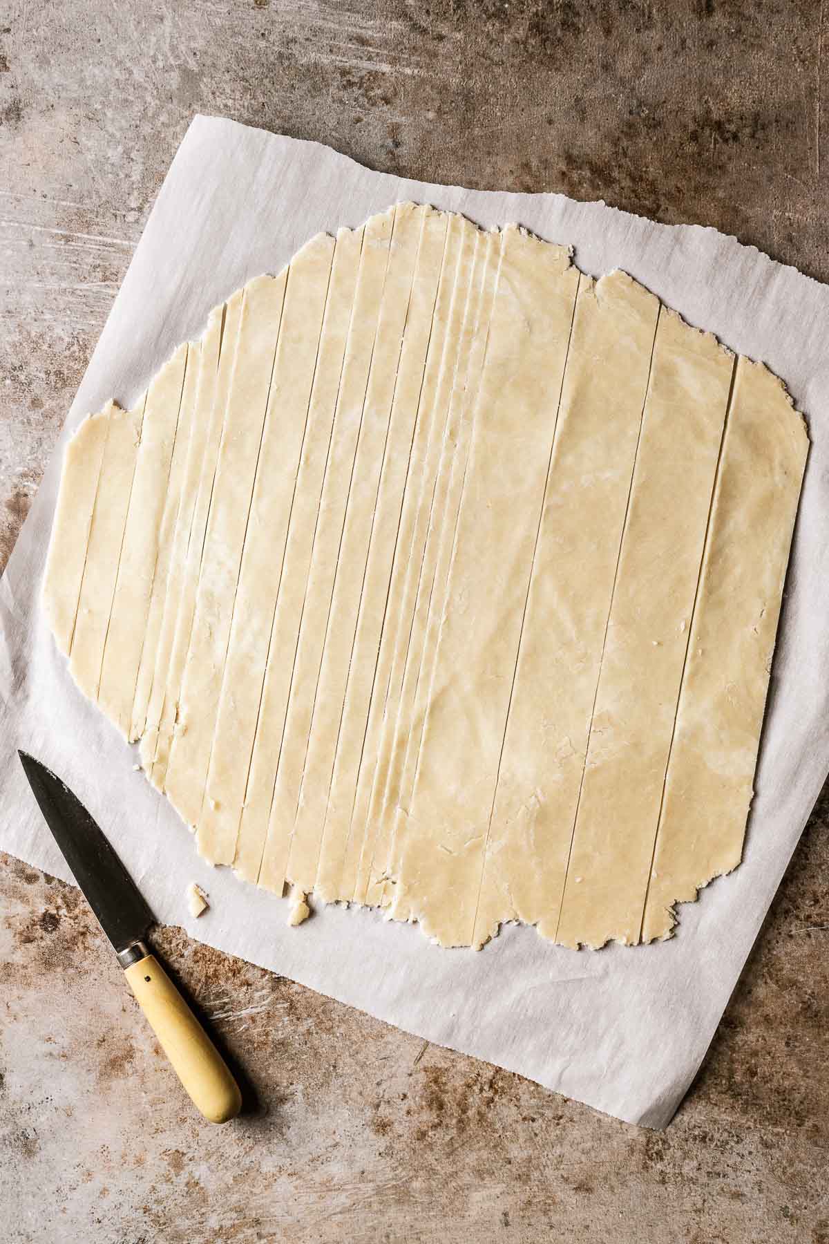 A rolled out sheet of pie dough on white parchment paper. The dough has random widths of lattice cut out and a knife rests nearby.