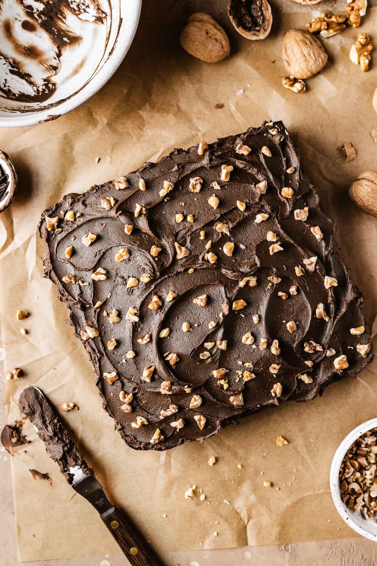 A chocolate walnut cake on brown parchment paper surrounded by white ceramic prep bowls, cracked walnuts and a ganache coated spatula.