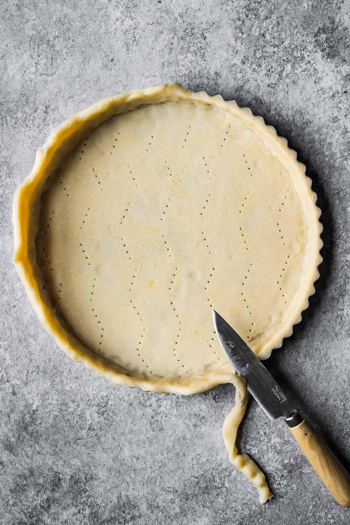 Pate sucree dough in a round tart pan with a knife being used to trim the edge of the dough flush with the pan.