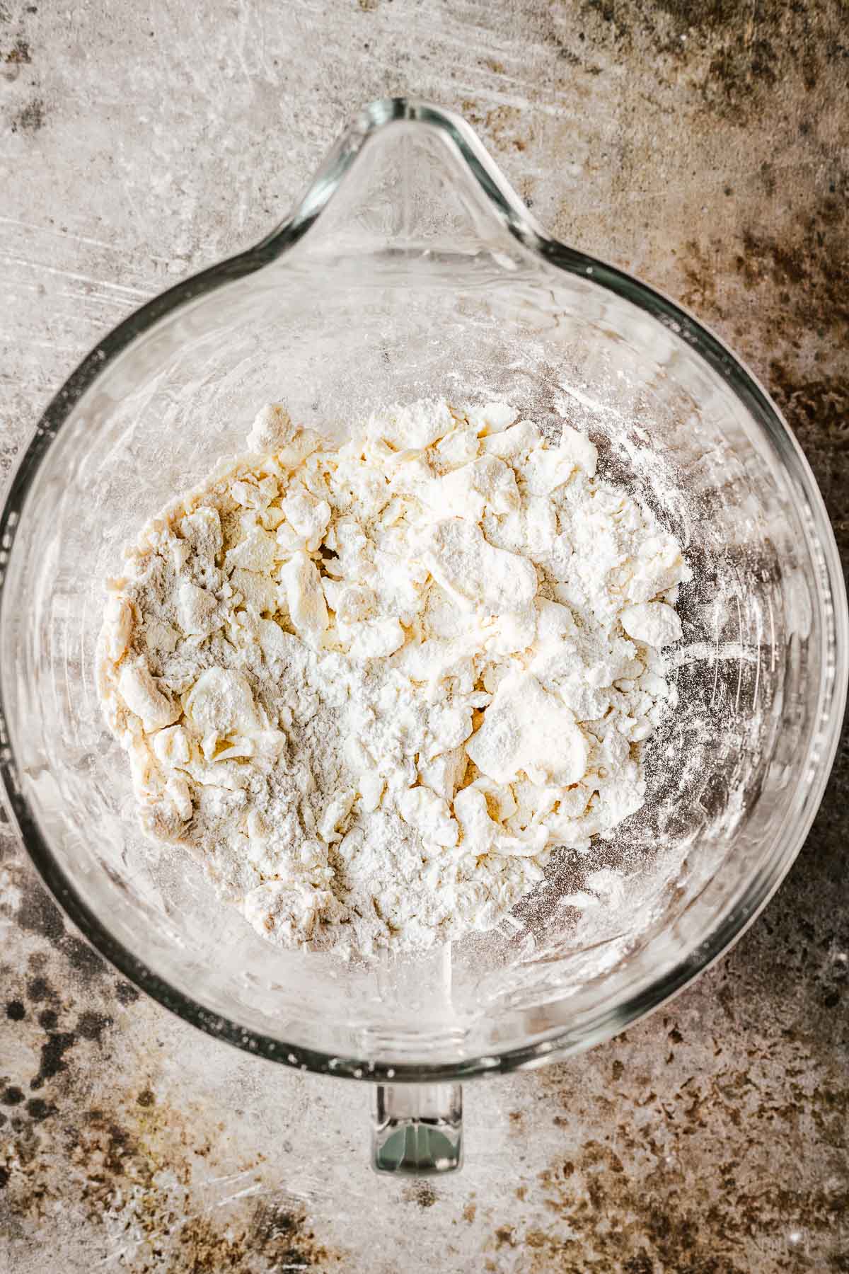 A glass mixing bowl filled with flour and salt with butter cubes flattened into shingle like pieces.