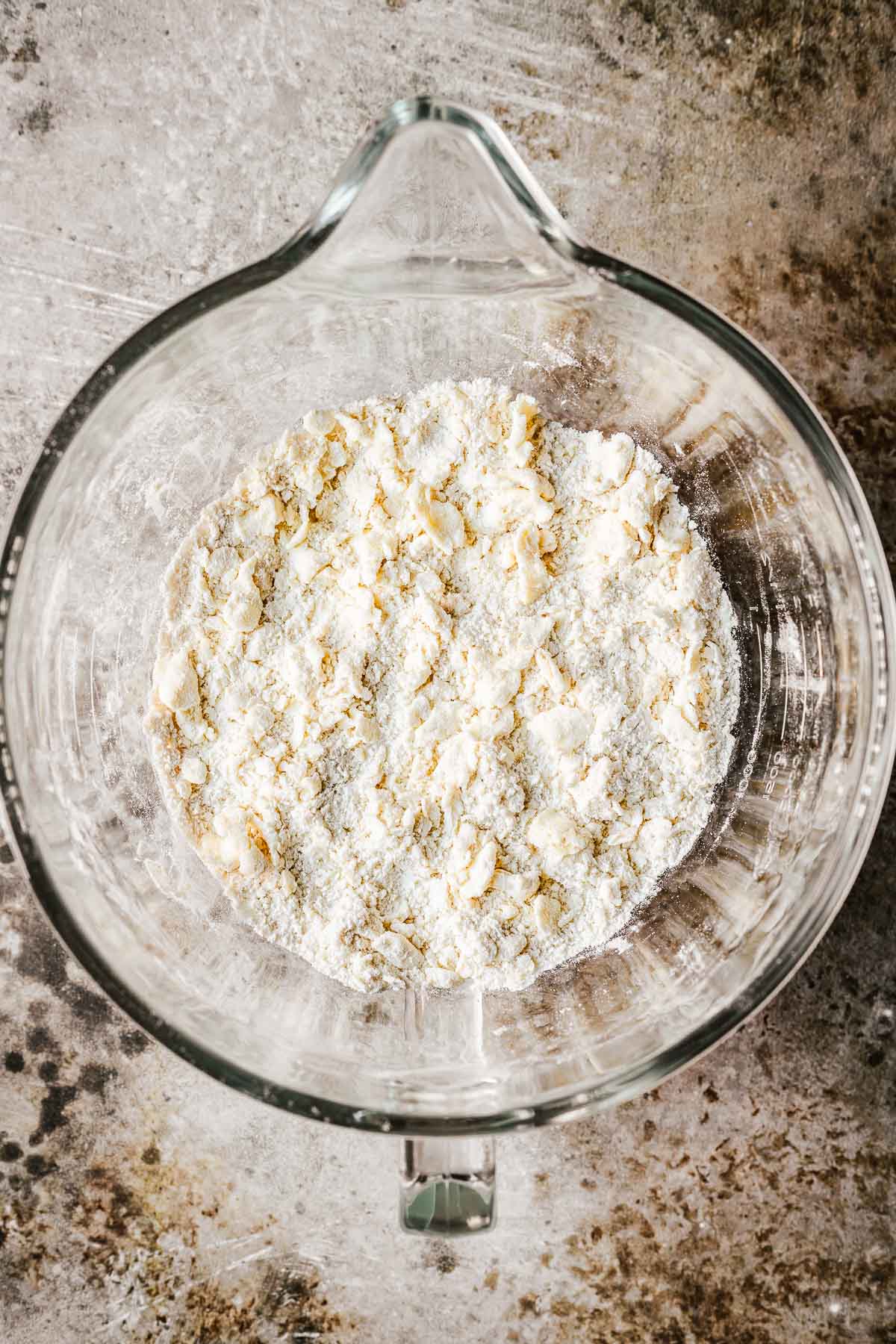 A glass mixing bowl with butter that has been worked into flour until it resembles small walnut halves.