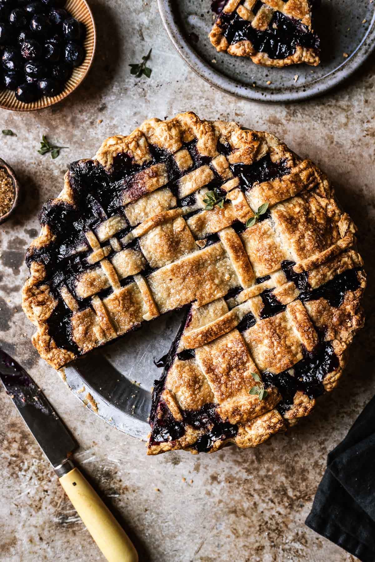 A golden brown, lattice topped blueberry pie with a single slice cut out and placed on a grey ceramic plate next to the pie. A knife rests nearby at bottom left.