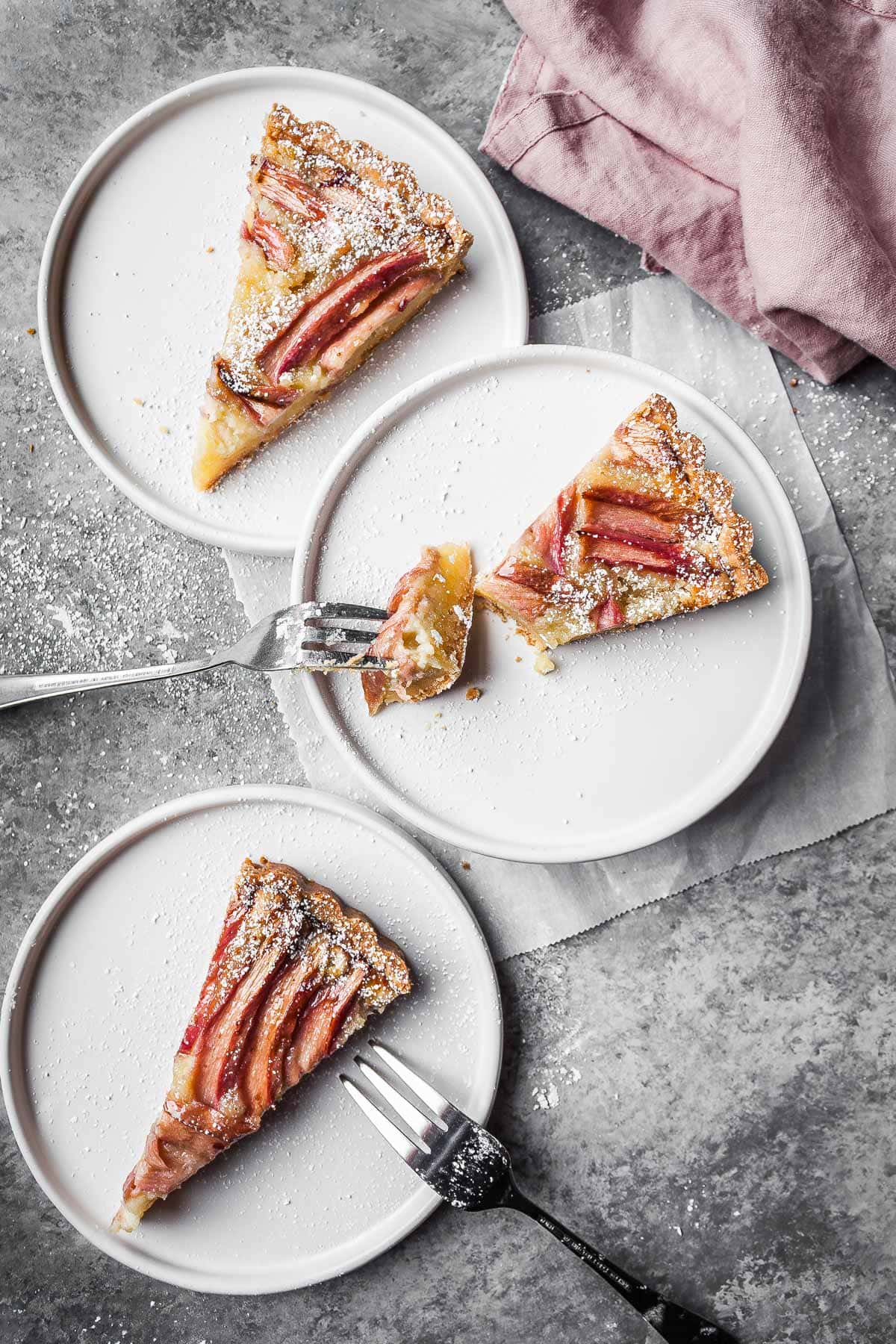 Three slices of fruit tart on white ceramic plates with two forks resting on the plates and a pink napkin nearby.