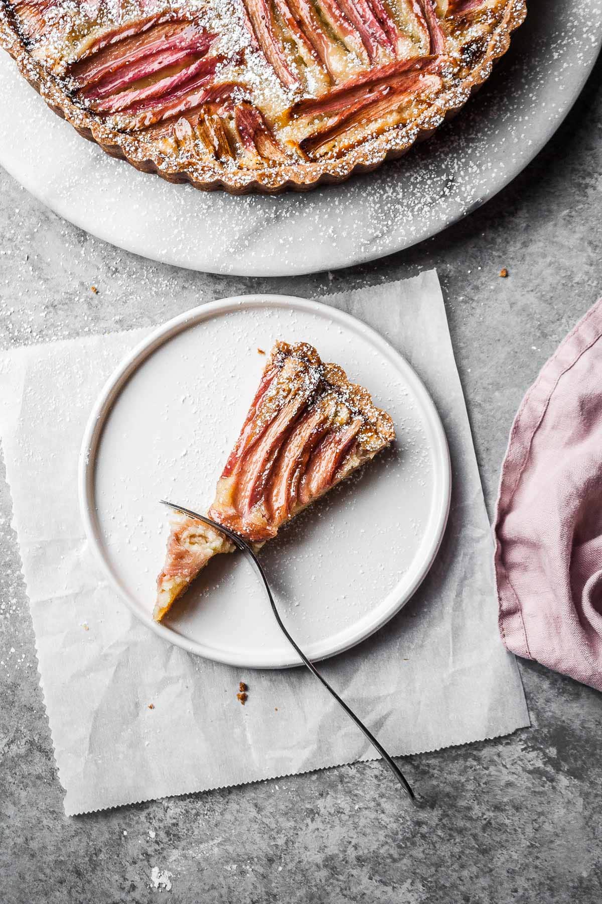 A slice of fruit tart on a white ceramic plate with a square of white parchment paper underneath. The rest of the tart peeks into the frame at top.