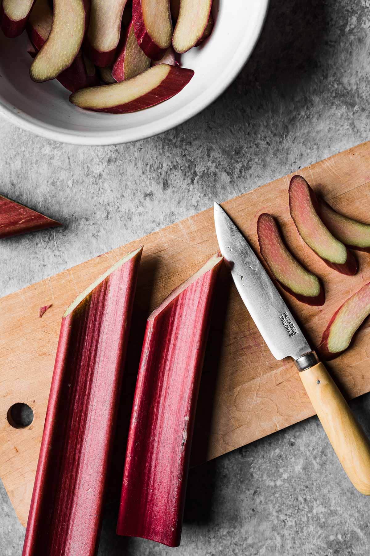 A wooden cutting board with reddish pink rhubarb stalks cut on the bias. A wooden handled paring knife rests nearby.