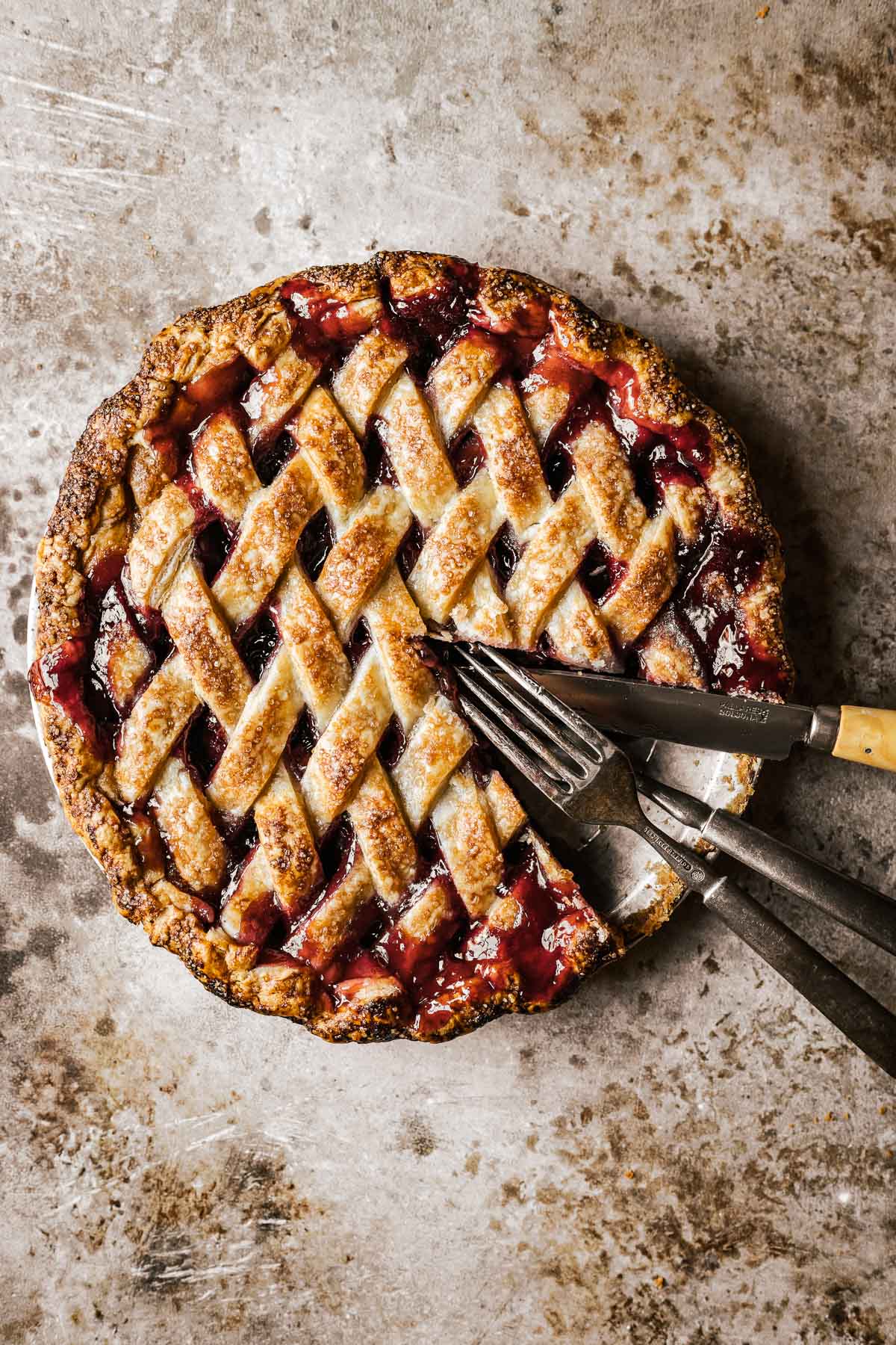 A diamond lattice topped pie with a slice removed. In the place where the slice was rest two forks and a knife.