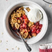 A white ceramic bowl filled with a serving of strawberry apple crisp and a scoop of vanilla ice cream. A spoon rests in the crisp.