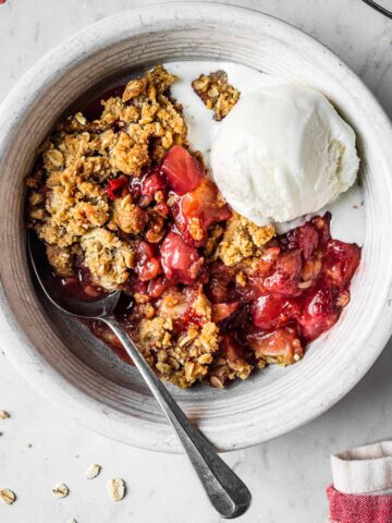 A white ceramic bowl filled with a serving of strawberry apple crisp and a scoop of vanilla ice cream. A spoon rests in the crisp.