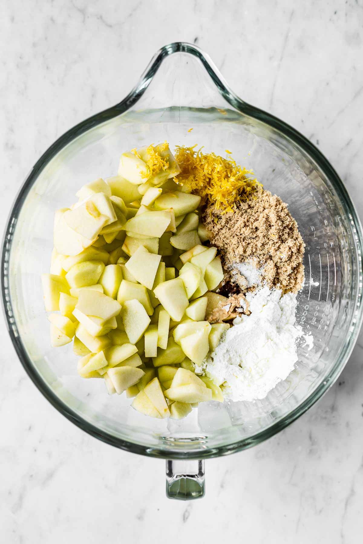 A large glass mixing bowl filled with apples, lemon zest, brown sugar and cornstarch for a fruit crisp.