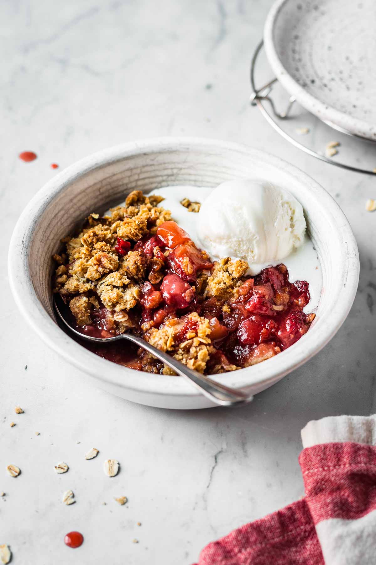 A white ceramic bowl filled with a serving of strawberry apple crisp. The crisp is topped with a scoop of vanilla ice cream, which is beginning to melt at the bottom.