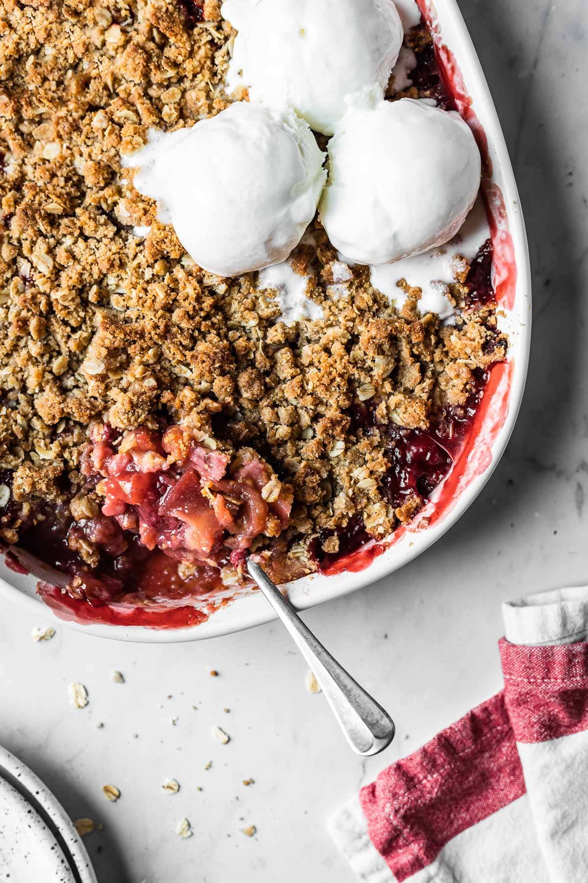 A white ceramic baking dish with a baked strawberry apple crisp topped with three scoops of vanilla ice cream. A spoon scooped into the crisp rests on the side of the dish.