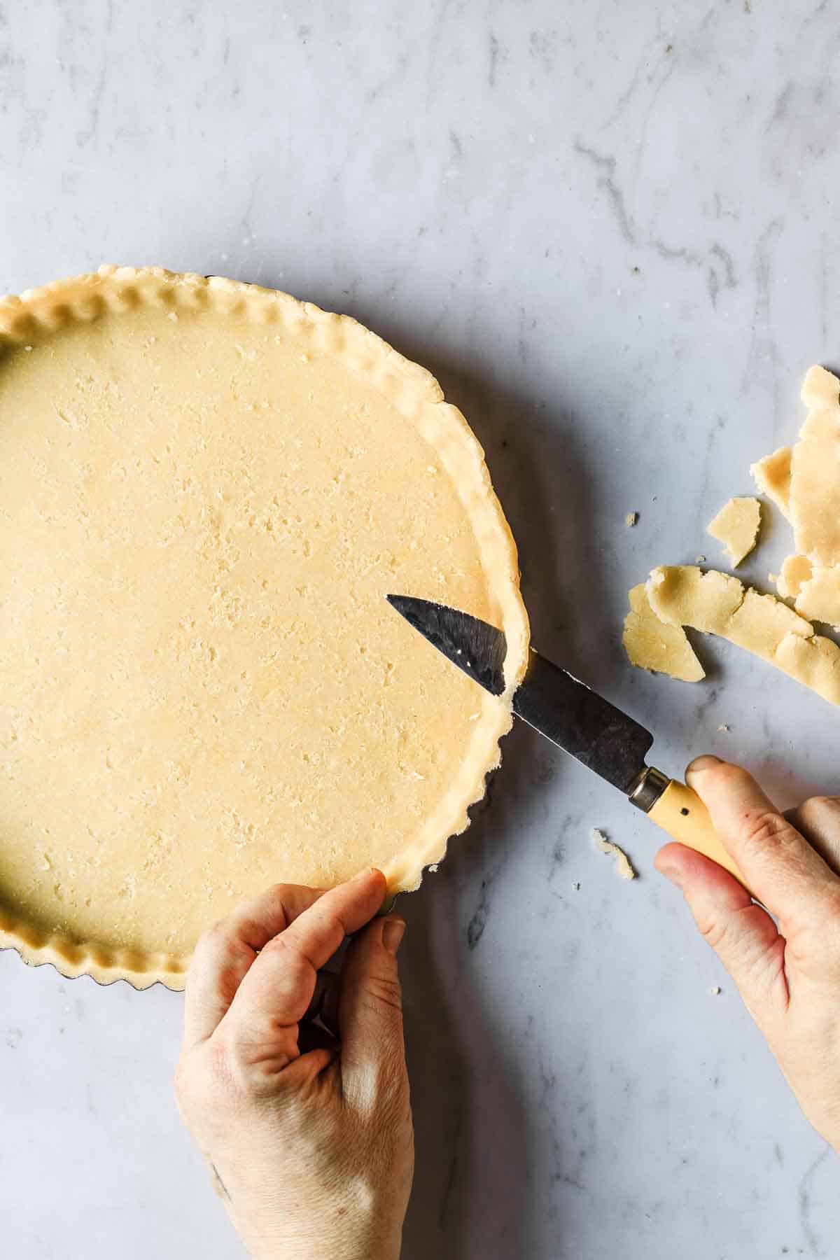 Hands holding a knife and trimming extra dough off tart.