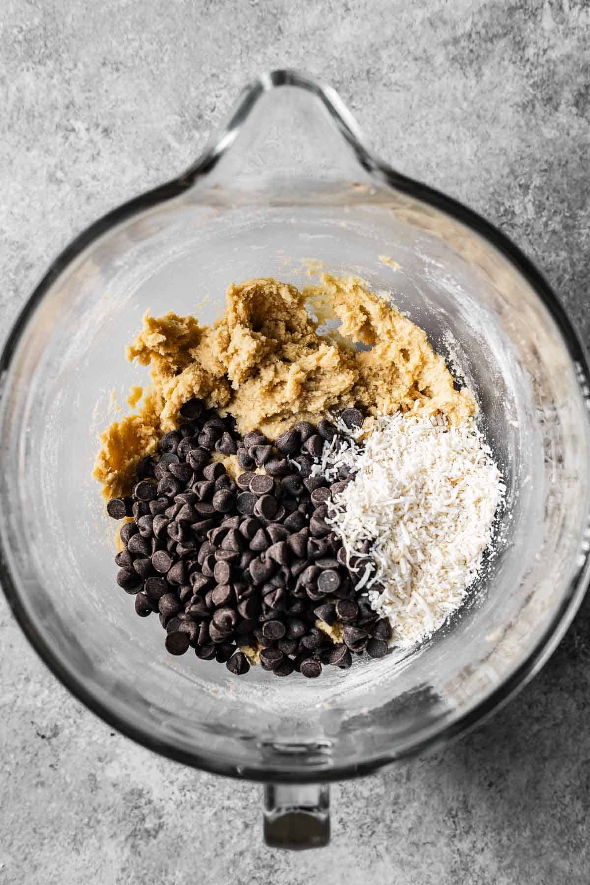 A mixing bowl with chocolate chips and shredded coconut on top of cookie dough.