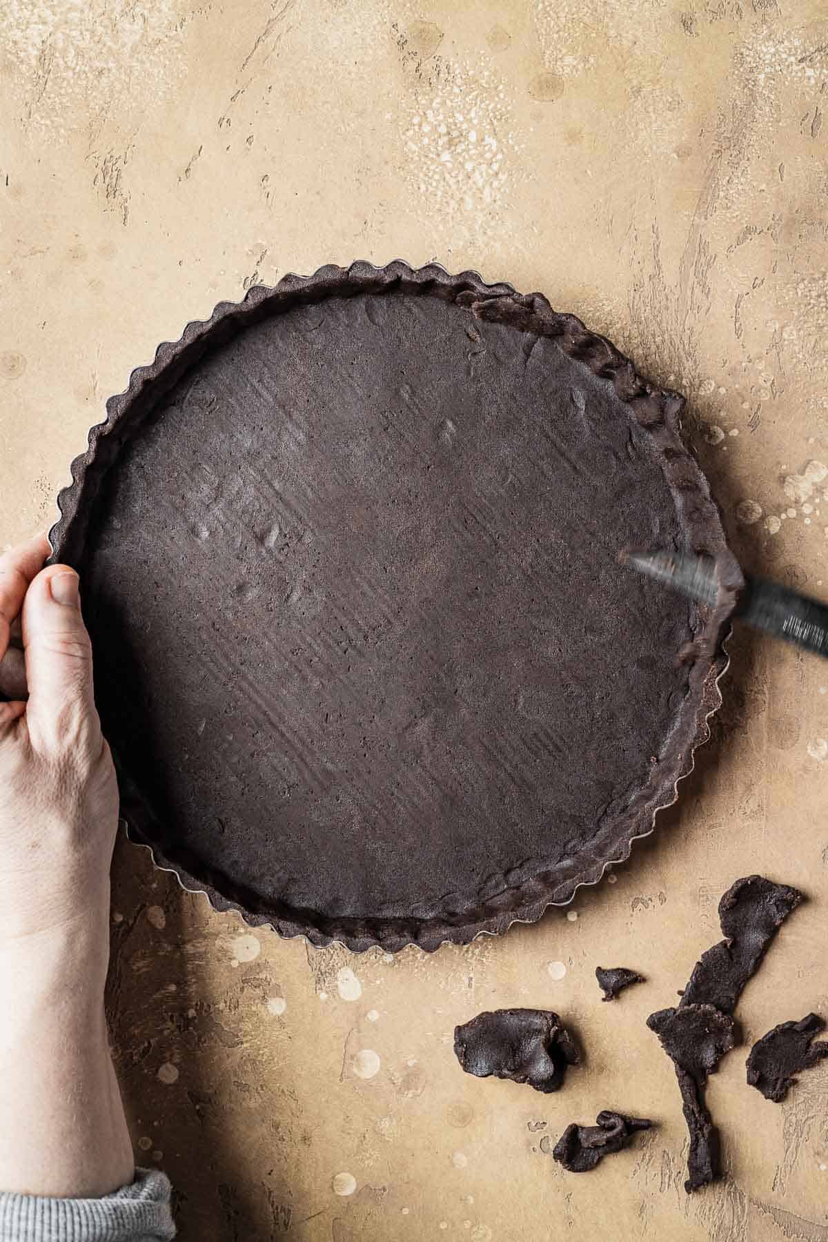 Hands trimming the edge of an unbaked tart shell with a knife.