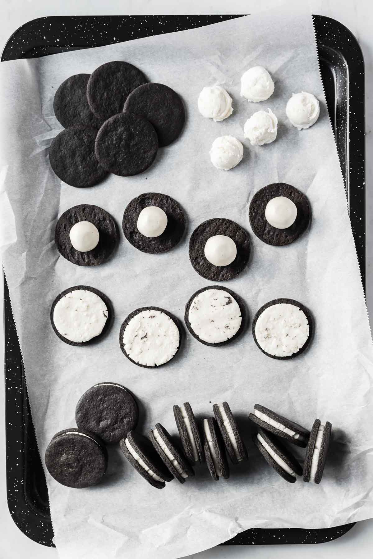 Homemade Oreo cookies in different stages of assembly on a black baking sheet.