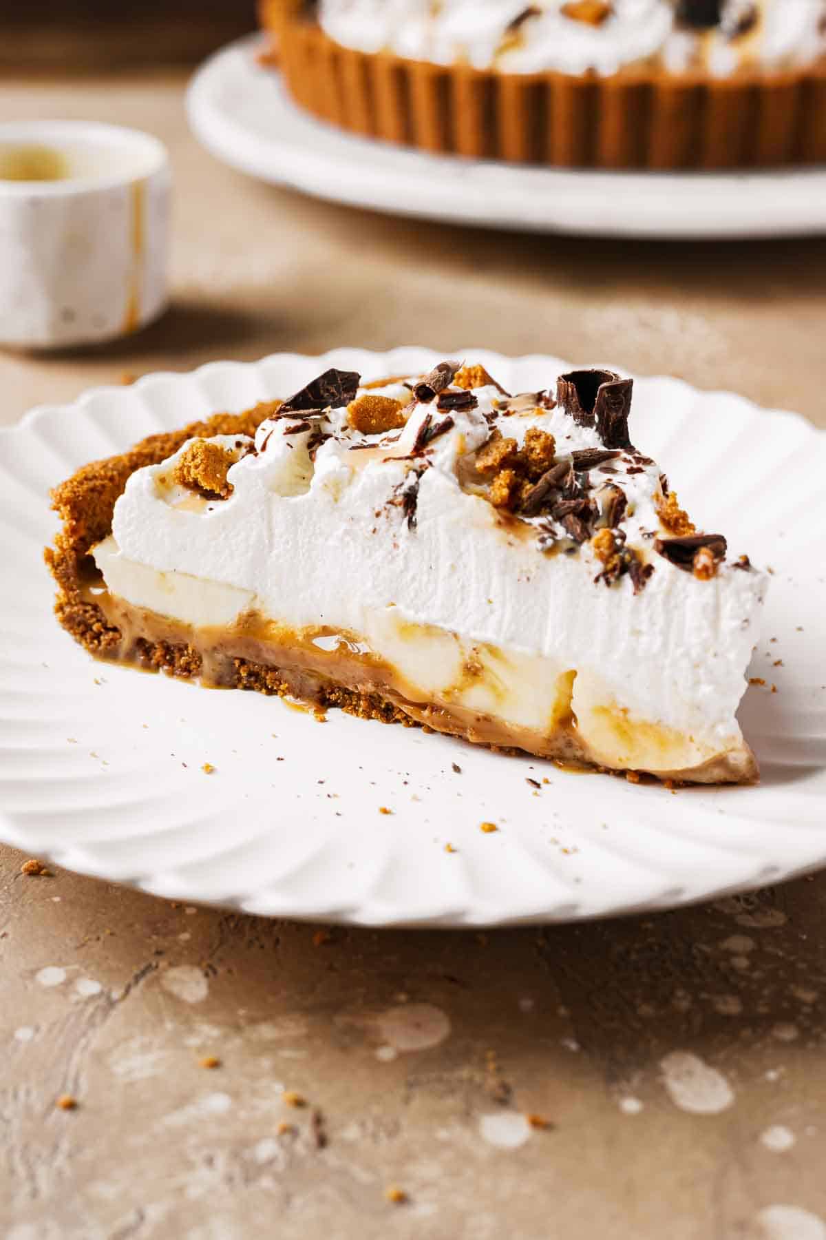 A slice of banoffee tart on a plate showing layers of dulce de leche, bananas and whipped cream.