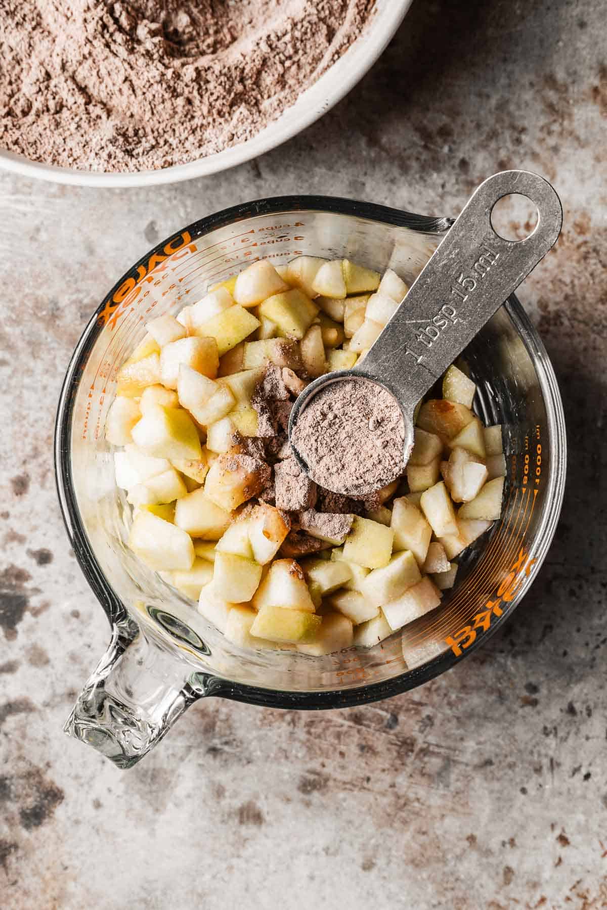 A glass measuring cup with diced pears and a tablespoon of flour mixture on top.