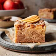 A slice of moist apple cake on a ceramic plate with three thin slices of fresh apple on top.