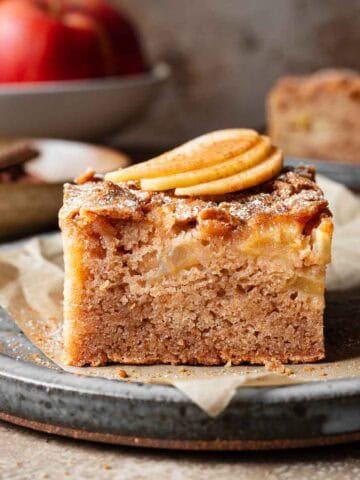 A slice of moist apple cake on a ceramic plate with three thin slices of fresh apple on top.