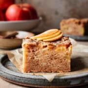 A slice of moist apple cake on a ceramic plate with three thin slices of fresh apple on top.