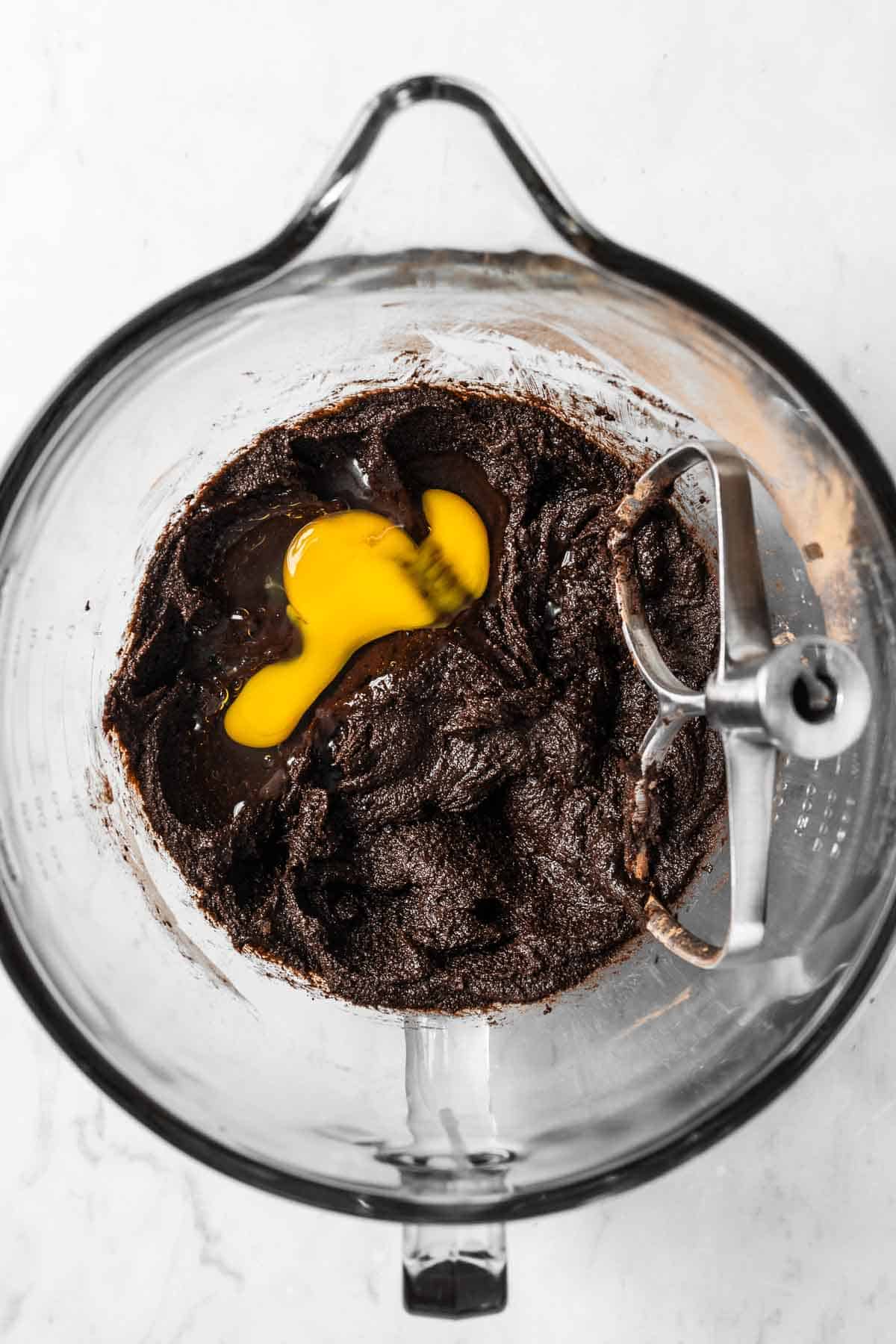 An egg added to melted butter, sugar and cocoa powder in a glass mixing bowl.