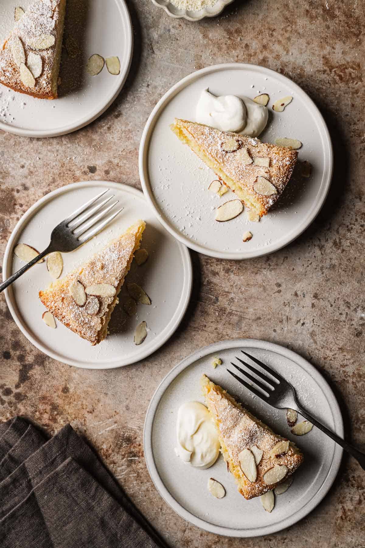 Slices of almond cake with slivered almonds and whipped cream on white ceramic plates.