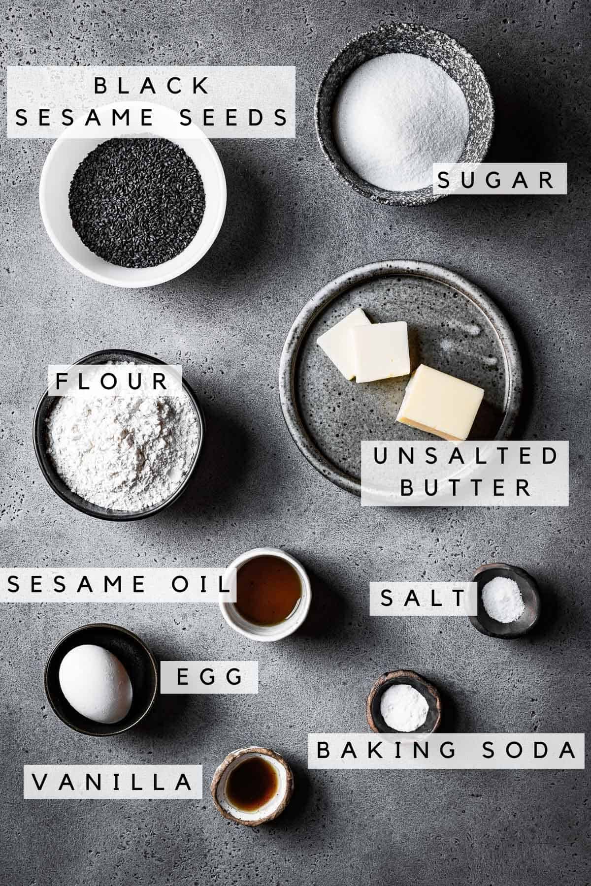 Labeled cookie ingredients in bowls on a grey stone surface.