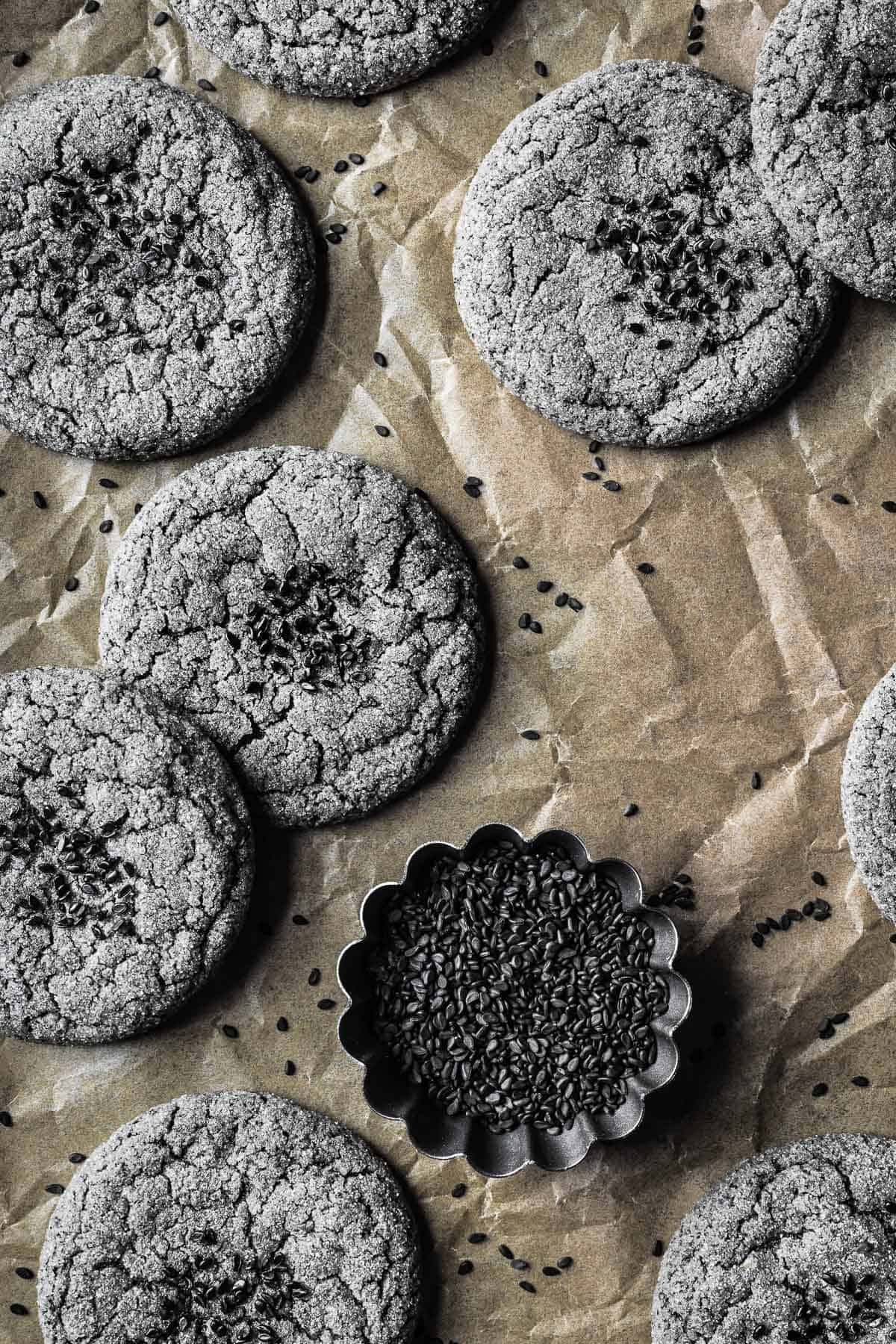 Black sesame cookies on brown parchment paper with a bowl of black sesame seeds nearby.