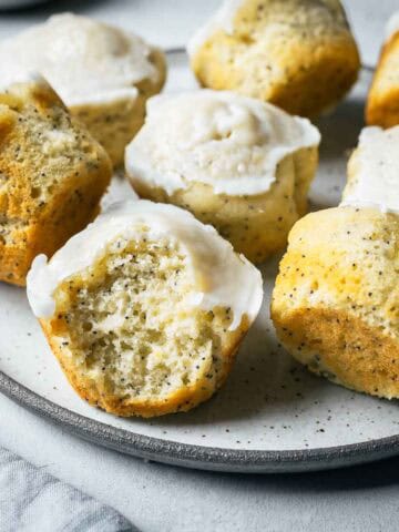Lemon poppy seed muffins on a ceramic plate with a large bite taken out of a muffin in the foreground, showing the crumb and poppy seeds inside.