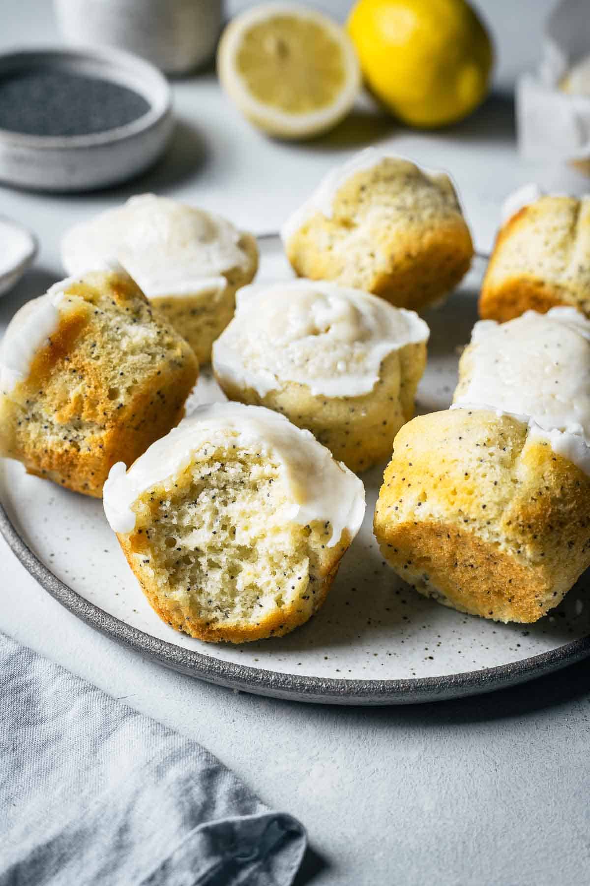Lemon poppy seed muffins on a ceramic plate with a large bite taken out of a muffin in the foreground, showing the crumb and poppy seeds inside.