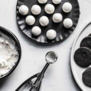 Scoops of Oreo creme filling on a black plate near a bowl of cream filling, measuring scoop, and plate of cookies.