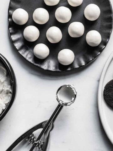 Scoops of Oreo creme filling on a black plate near a bowl of cream filling, measuring scoop, and plate of cookies.