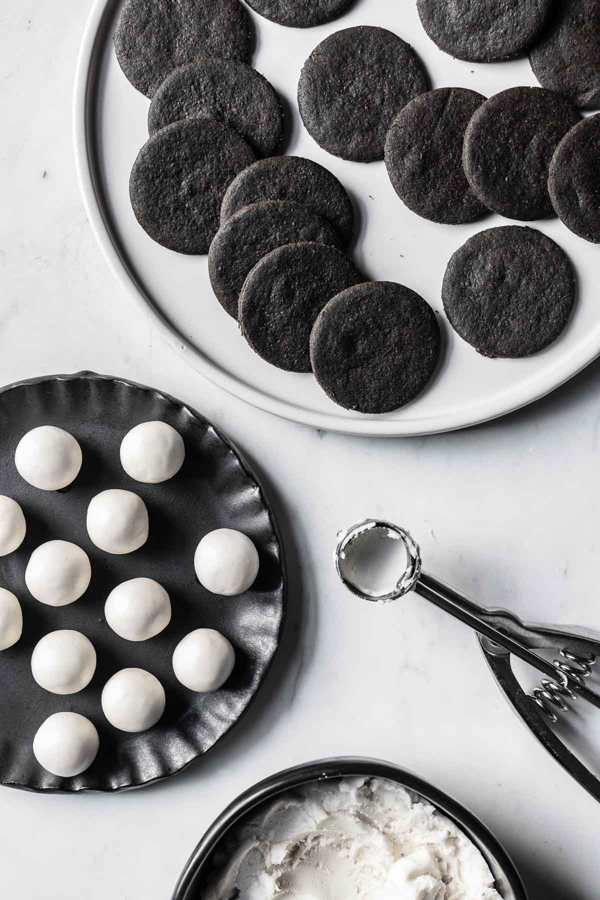 Scoops of Oreo creme filling on a black plate near a bowl of cream filling, measuring scoop, and plate of cookies.