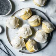 Lemon poppy seed muffins on a ceramic plate surrounded by lemons, a bowl of poppy seeds, a juicer and a pale blue linen napkin.