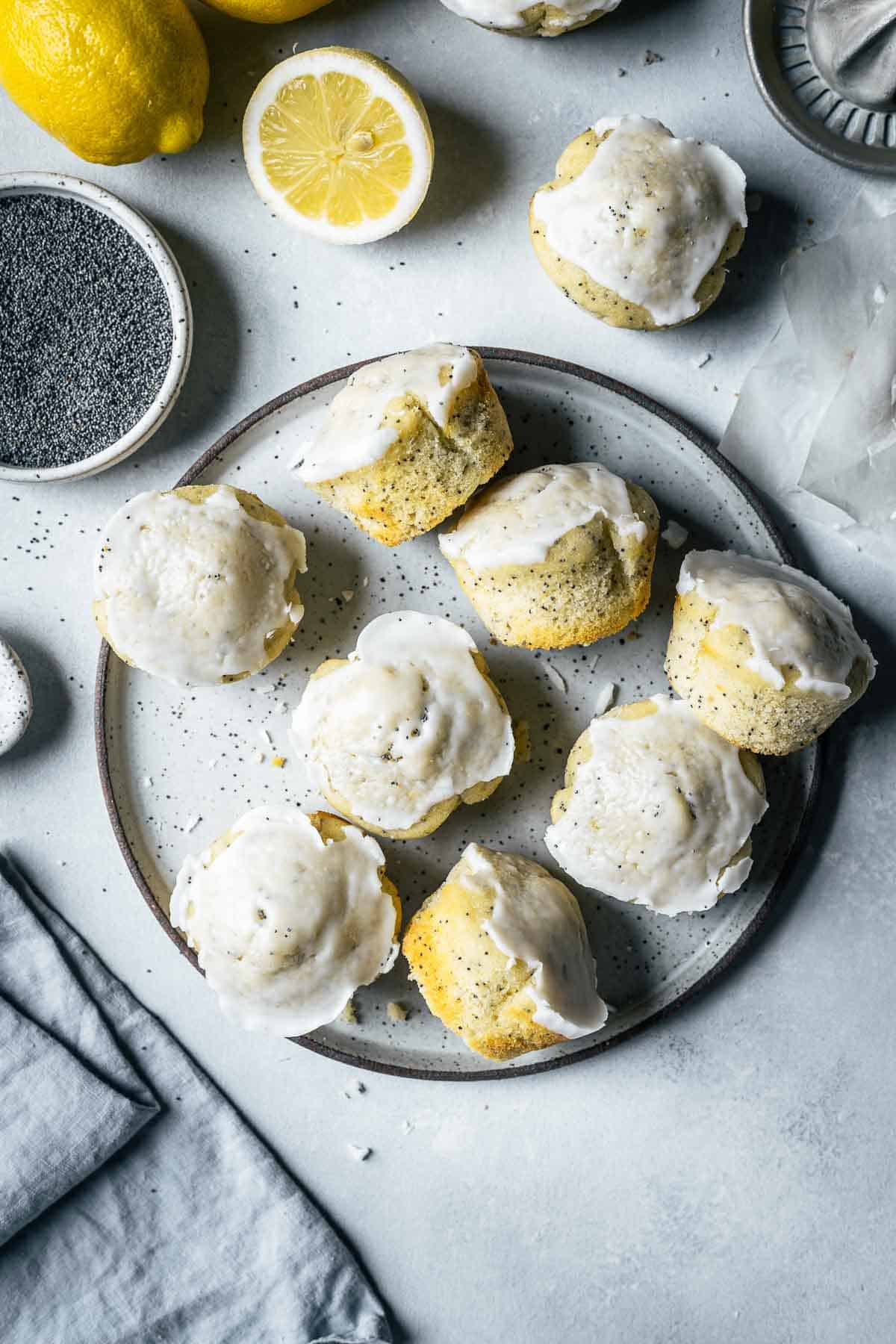 Lemon poppy seed muffins on a ceramic plate surrounded by lemons, a bowl of poppy seeds, a juicer and a pale blue linen napkin.