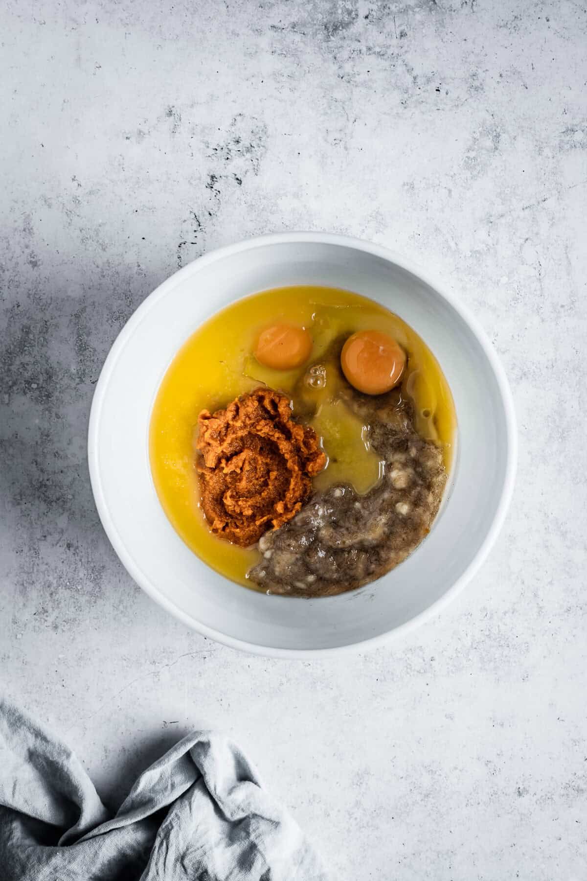 Eggs, melted butter, pumpkin and banana in a white glass mixing bowl.