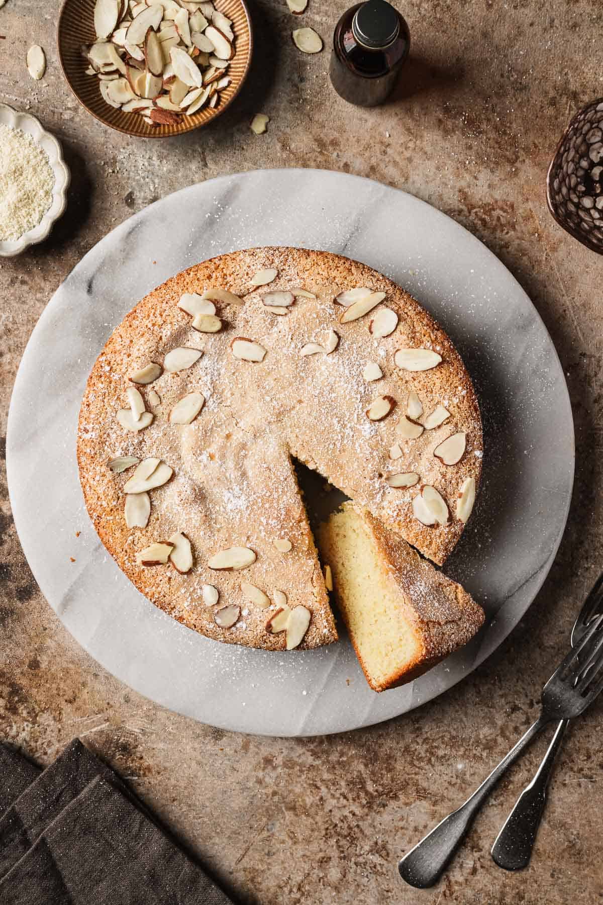 An almond cake on a marble platter with one slice cut and turned to show the crumb.