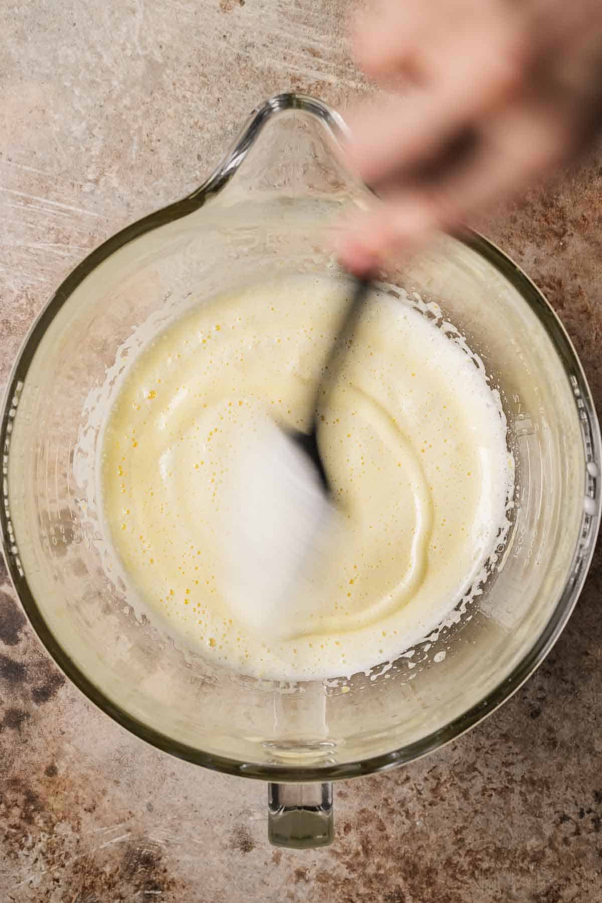 A hand holding a spatula over a mixing bowl, showing egg and sugar beaten to ribbon stage.