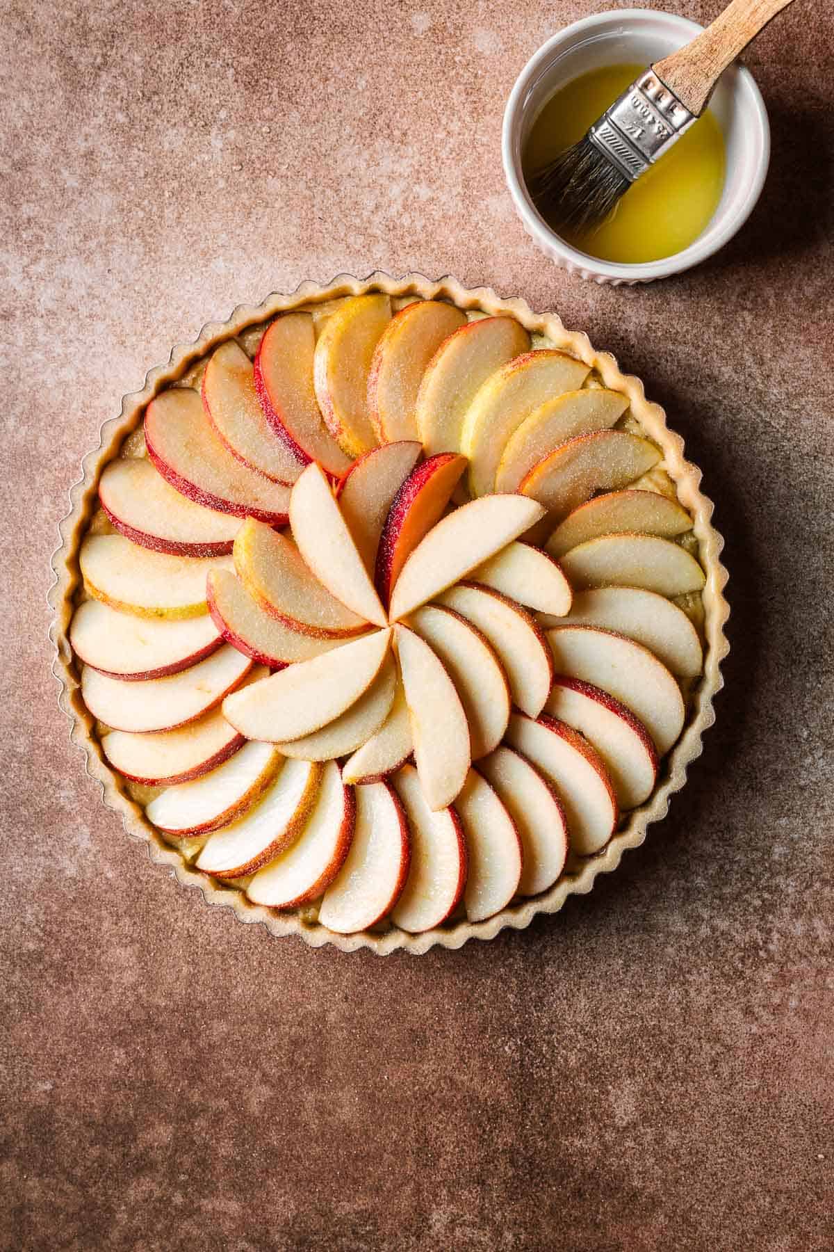 A round unbaked tart with sugar topped apple slices and a small bowl of melted butter with a pastry brush.